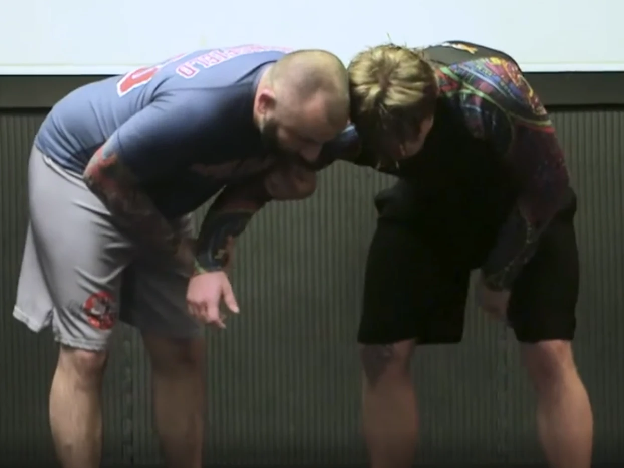 Two grapplers standing, top player has one arm of the defending player attached to his chest with both hands.